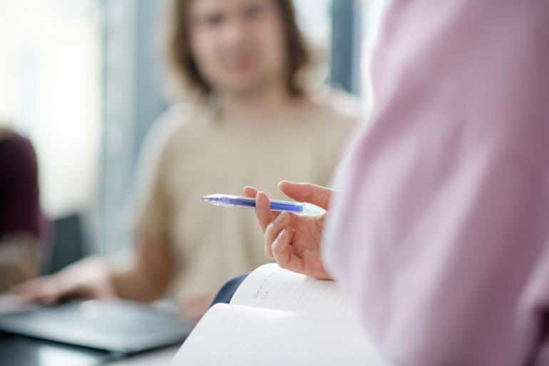 Someone is holding a pen and a notebook and talking to a blurred person in front of them.