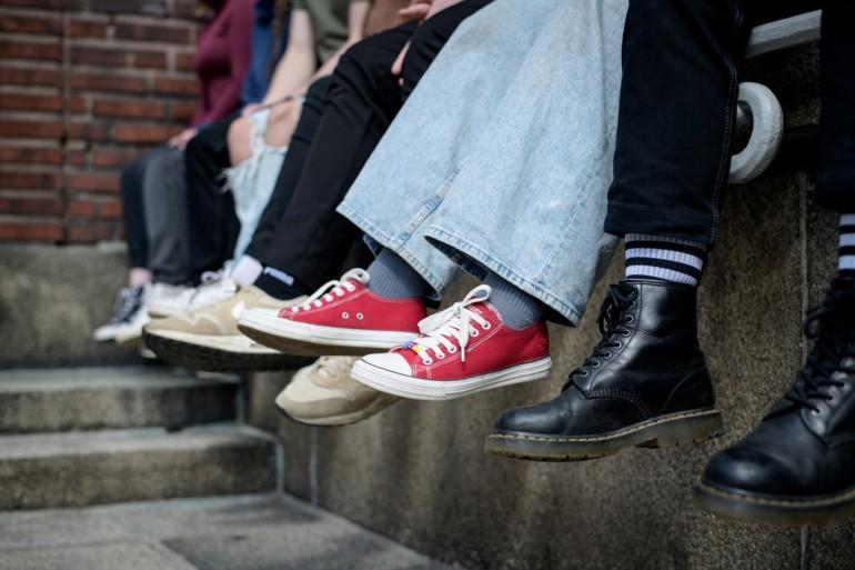 Kuvassa näkyy reunuksella vierekkäin rivissä istuvien nuorten jalkoja ja erilaisia kenkiä