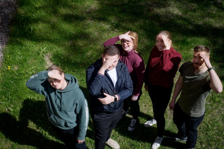 viisi nuorta seisoo nurmikolla ja katsoo ylöspäin kohti kameraa pitäen käsiä silmien edessä häikäisevän auringon valon takia