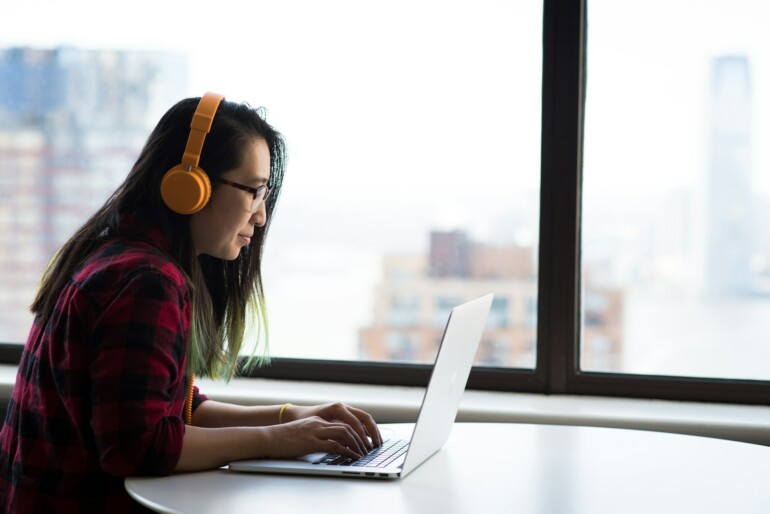 Nuori nainen istuu kuulokkeet päässä pöydällä kannettavan tietokoneen kanssa. Hän katsoo kannettavaa konettaan.