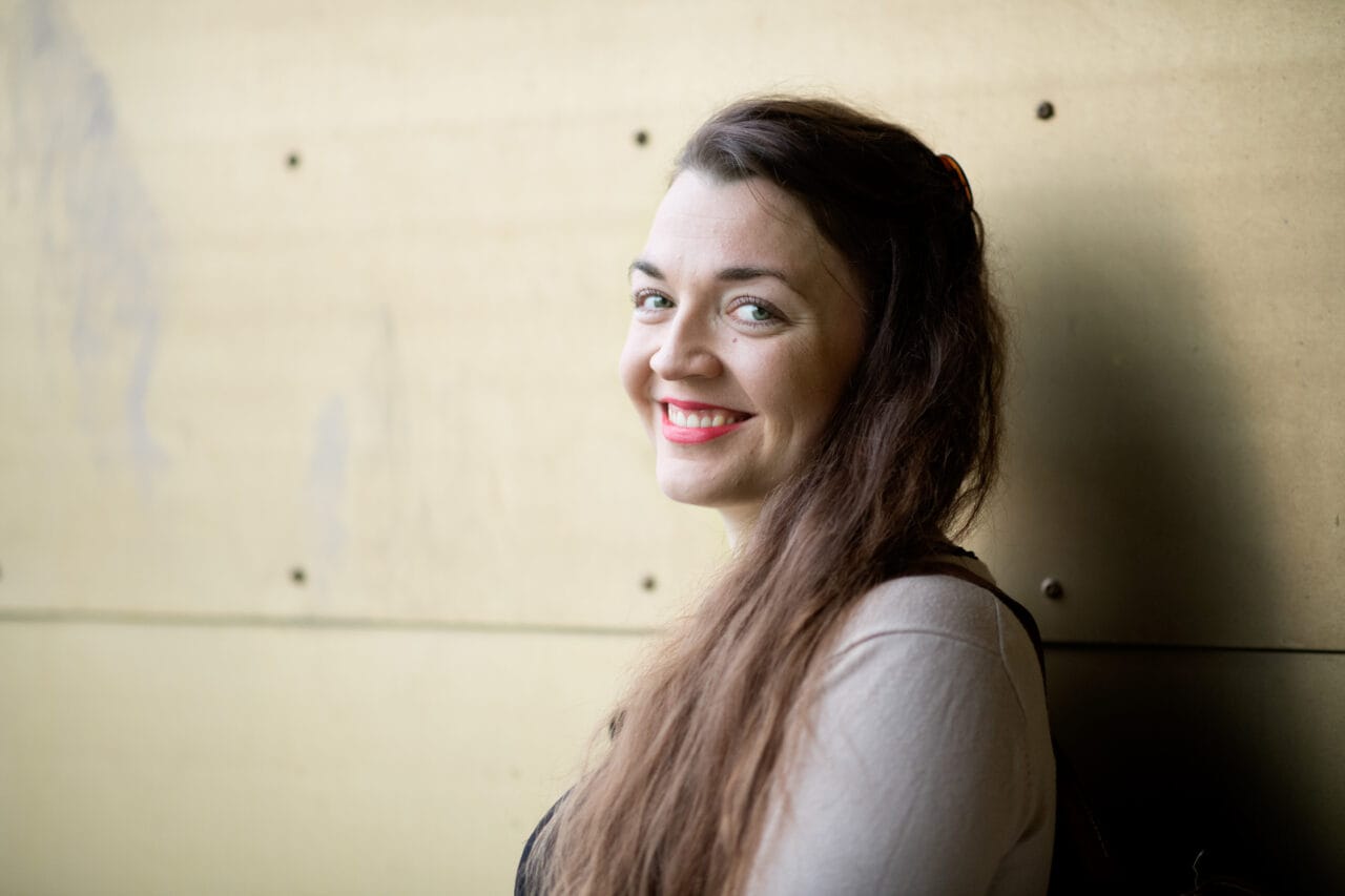 Pitkähiuksinen nainen seisoo seinän edessä hymyillen.