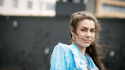 Nainen seisoo ulkona ja katsoo kameraan, yllään sininen romaninaisen puku, taustalla tumma seinä ja rakennuksia.