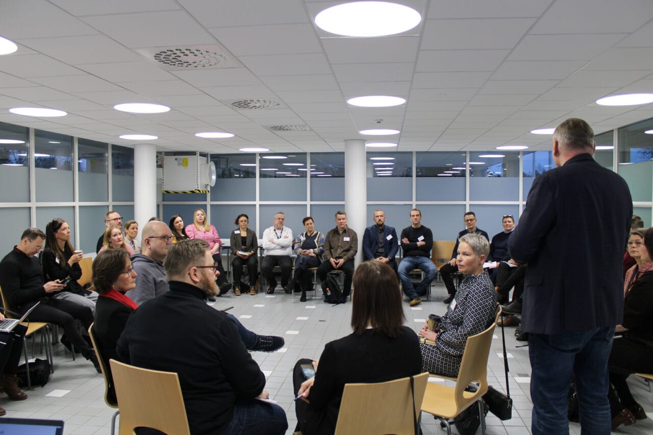 Suuri ryhmä 4Q-ammattikorkeakoulujen henkilöstöä istuu tuolirinkinä valoisassa kokoustilassa osallistuen yhteiseen keskusteluun. Yksi henkilö seisoo ja puhuu muille, kun taas muut kuuntelevat, tekevät muistiinpanoja tai pitävät taukovälineitä käsissään. Taustalla näkyvät suuret ikkunat ja vaalea sisustus.