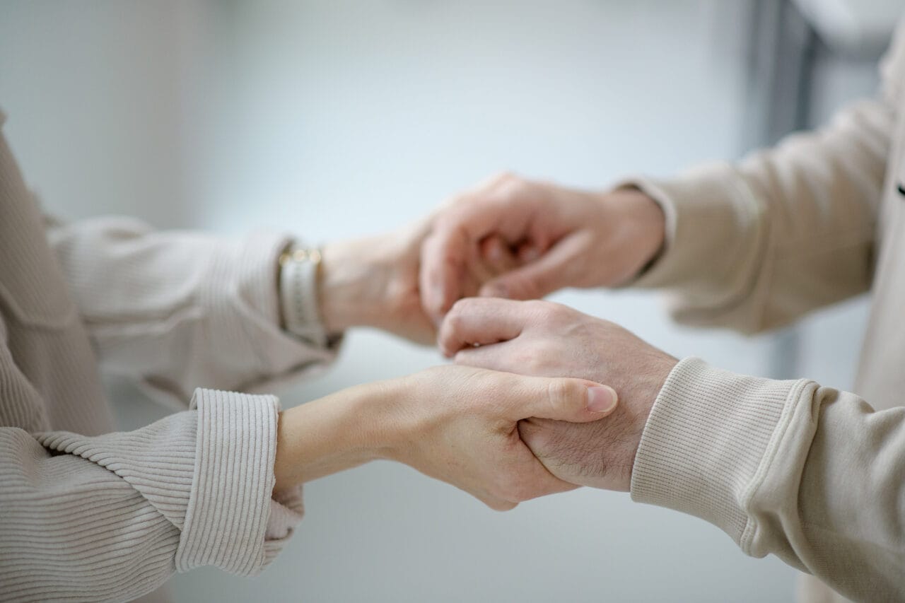 Kuvassa nuoremman ja iäkkäämmän henkilön kädet, jotka pitävät toisistaan kiinni. Tausta on vaalea.