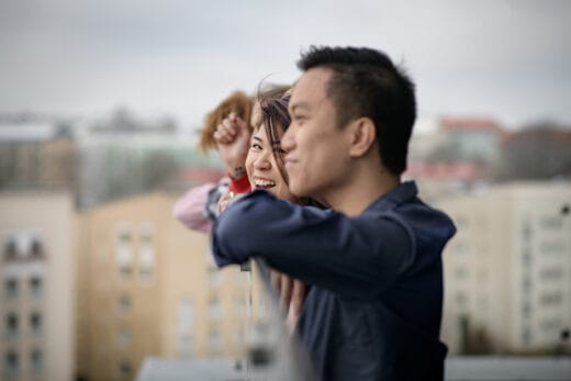 Nuoret nojaavat kaiteeseen ulkona. Kamera tarkentuu nuoreen joka katsoo etualalla olevaa toista nuorta nauraen.