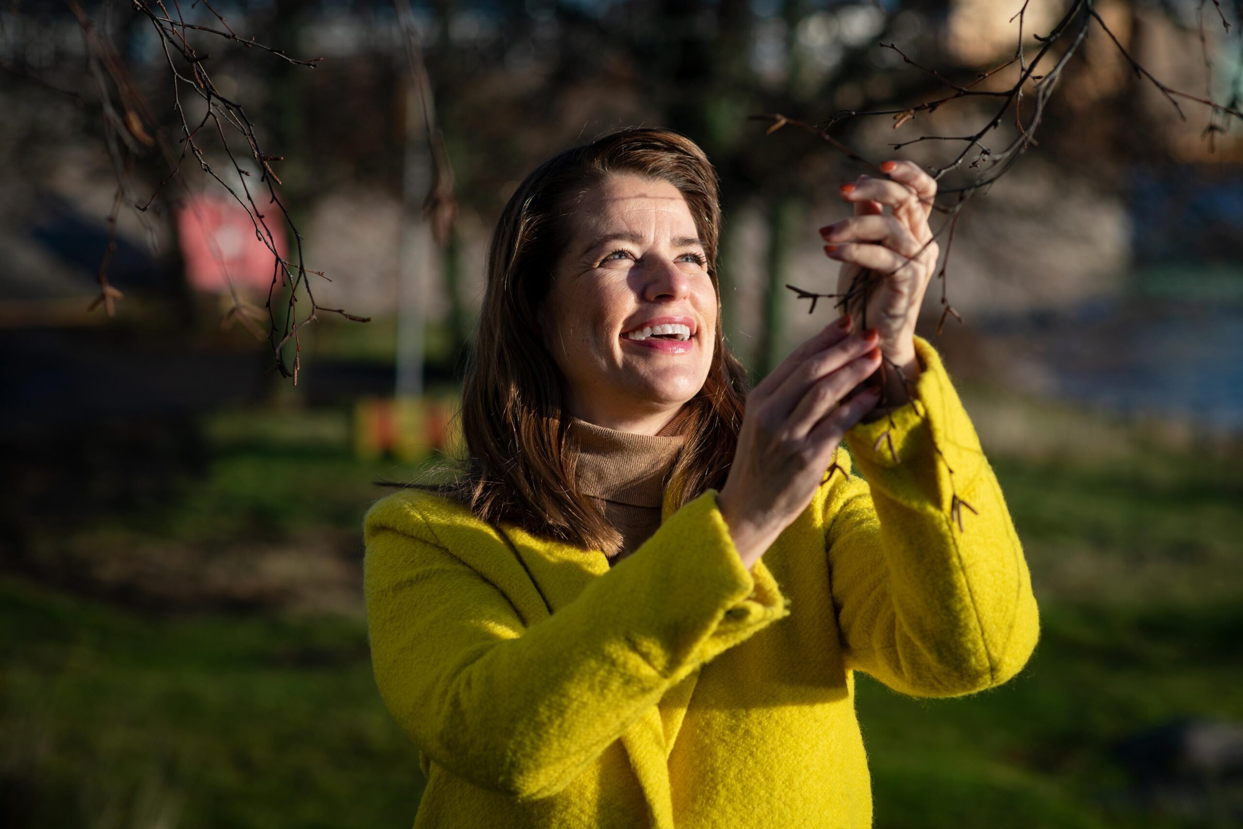 Marianne Heikkilä hymyilee auringonpaisteessa ja pitää käsiään puun oksilla.