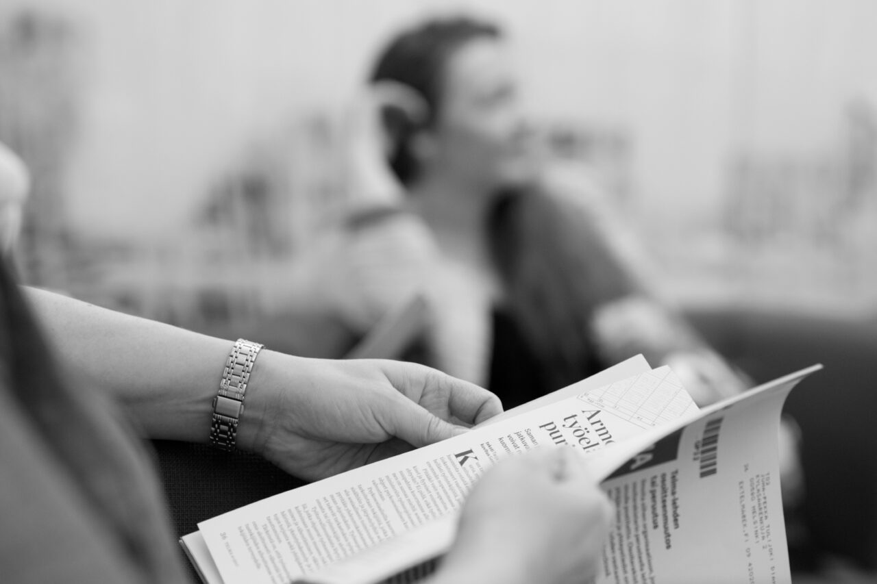 Etualalla henkilö lukee lehteä. Taustalla toinen sumuinen henkilö istuu sohvalla. Mustavalkoinen kuva.