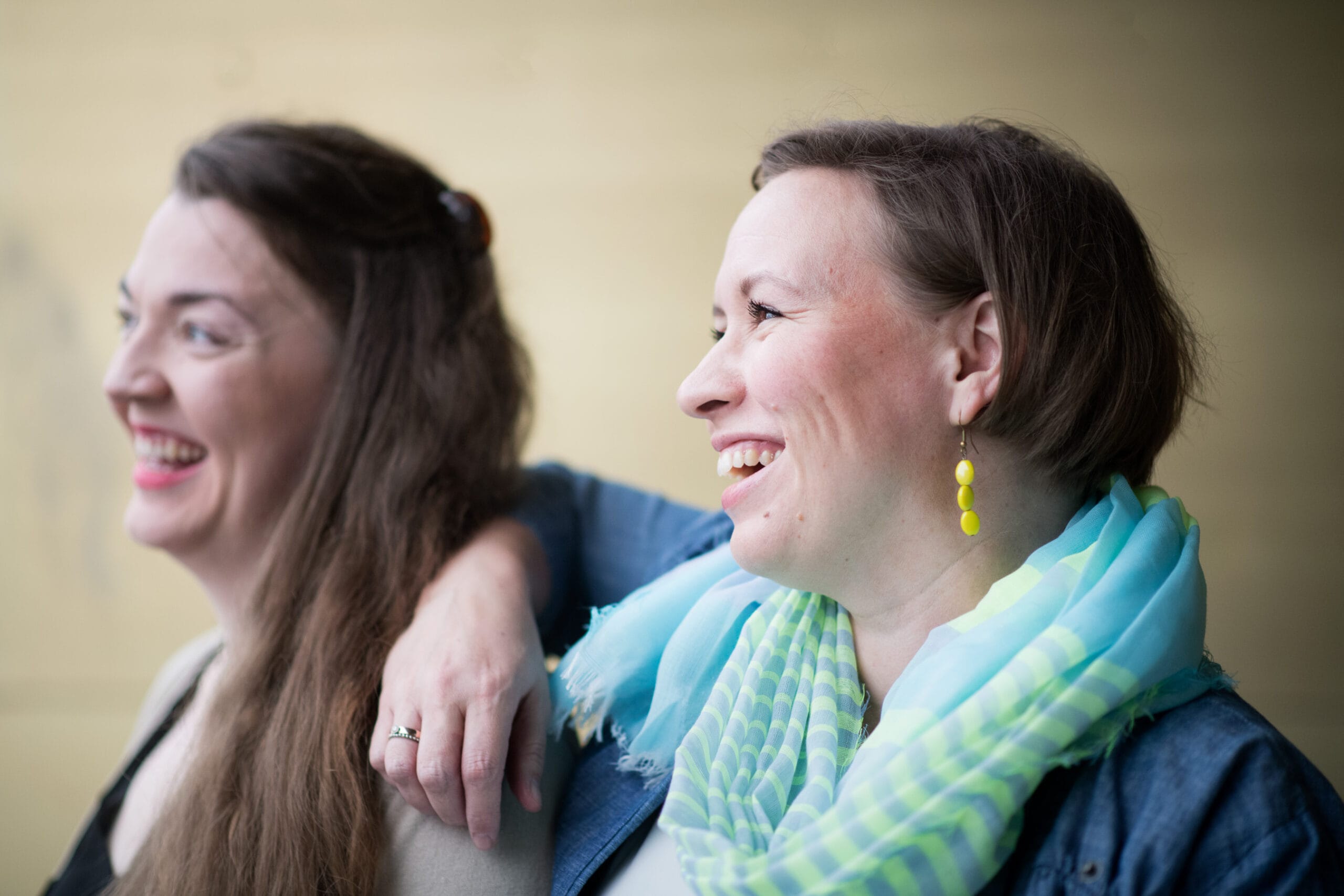 Kaksi naista vierekkäin, toinen nojaa kätensä toisen olalle. Naiset nauravat.