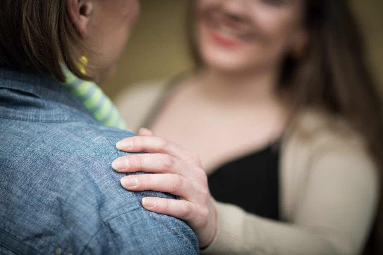 Kaksi naista. Kuva on kuvattu toisen naisen olan takaa. Toinen nainen pitää kättään naisen olkapäällä ja hymyilee.