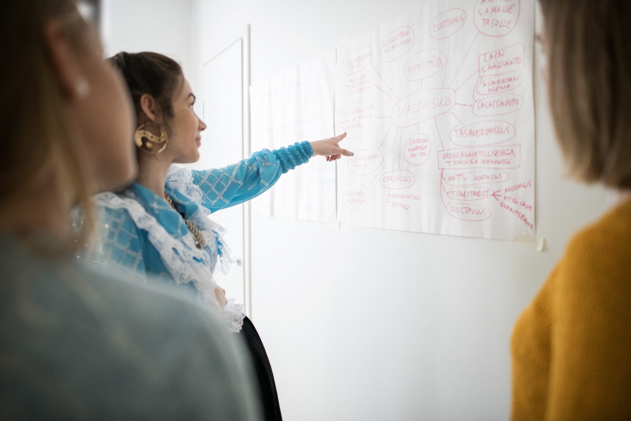 Romaninainen osoittaa seinälle ripustetulta paperilta tekstiä kahdelle vieressä seisovalle naiselle.