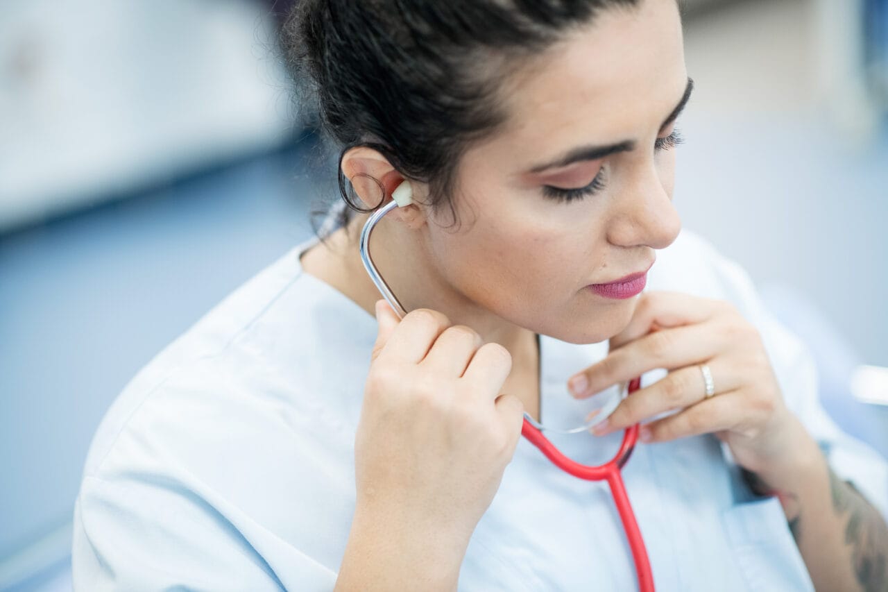 Kuvassa näkyy hoitaja yllään vaalea työasu. Hän asettaa stetoskoopin korvilleen.