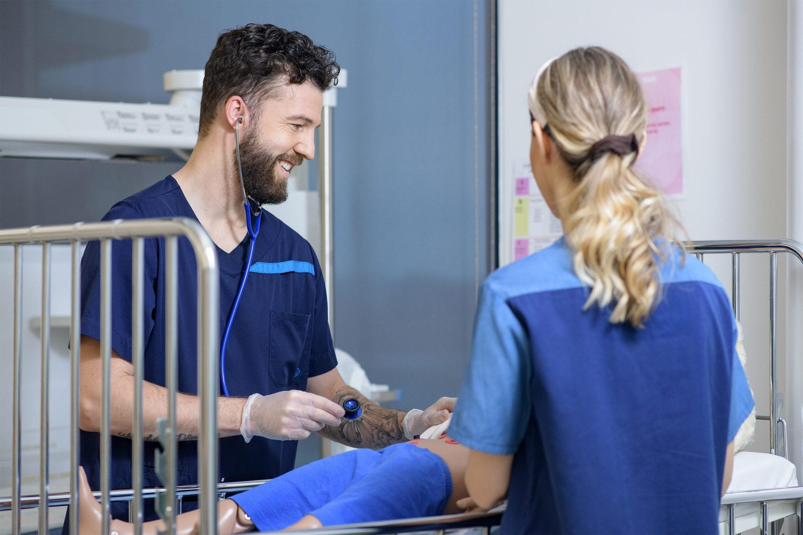 Kaksi sairaanhoitajaopiskelijaa hoitavat nukkepotilasta, joka makaa sairalasängyllä.