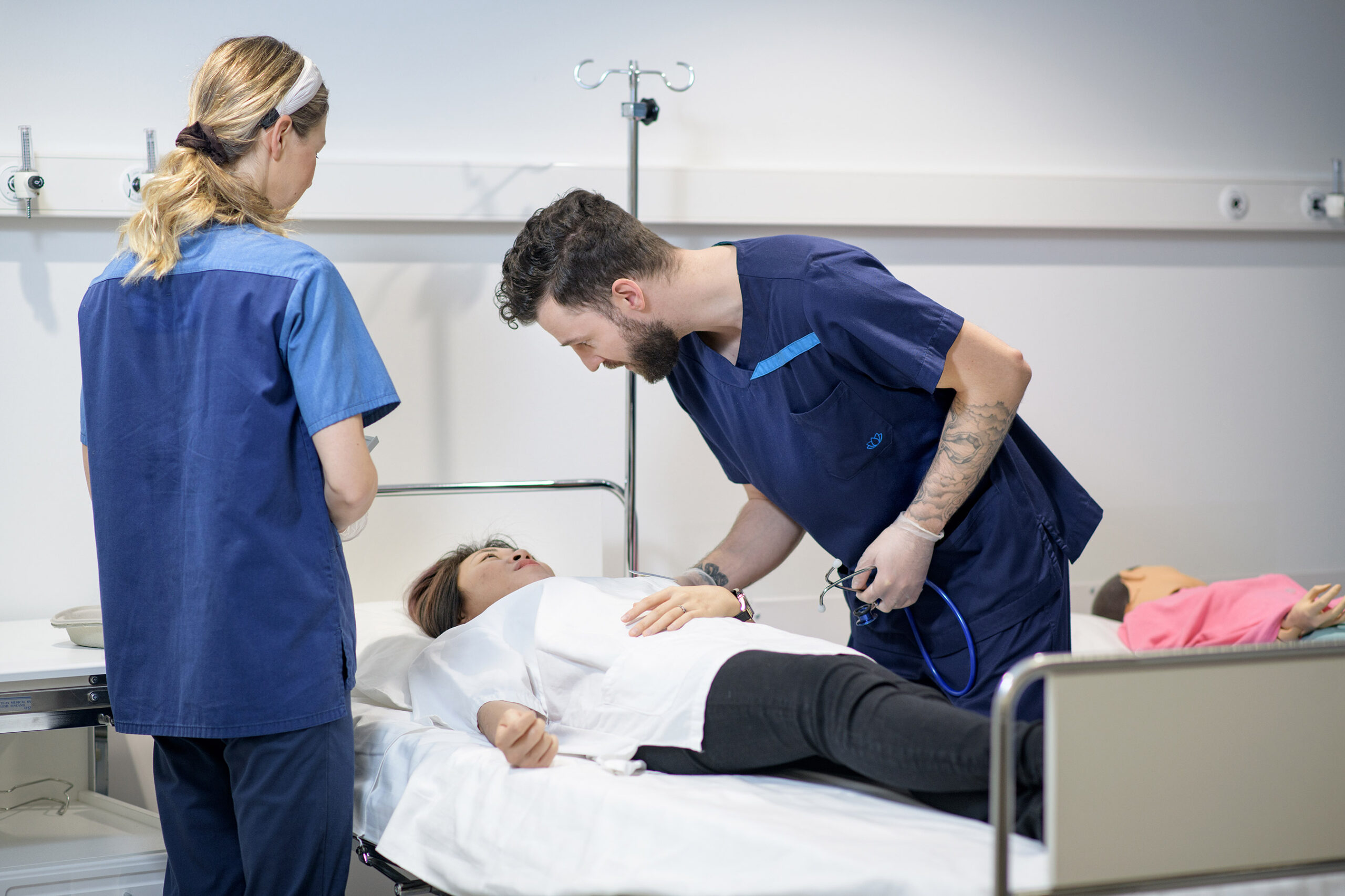 Kaksi sairaanhoitajaksi pukeutunutta opiskelijaa kumartuneena sängyssä makaavan simulaatiopotilaan puoleen, kuvituskuva.