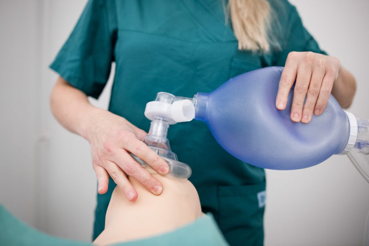 Henkilö, jolla on yllään vihreä hoitoasu antaa hengitysapua harjoitusnukelle. Hän pitelee sinistä käsipumppua ja painaa kasvomaskia nuken kasvoille.