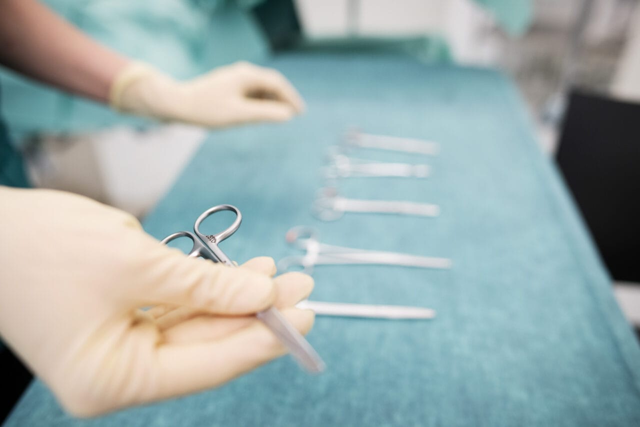 Kädet, joissa on vaaleat suojakäsineet. Etualalla käsi pitelee metallisia kirurgisia saksia. Taustalla pöydällä on rivi muita hoitovälineitä tai instrumentteja. Pöydän pinta on sinivihreä ja tausta on sumea.