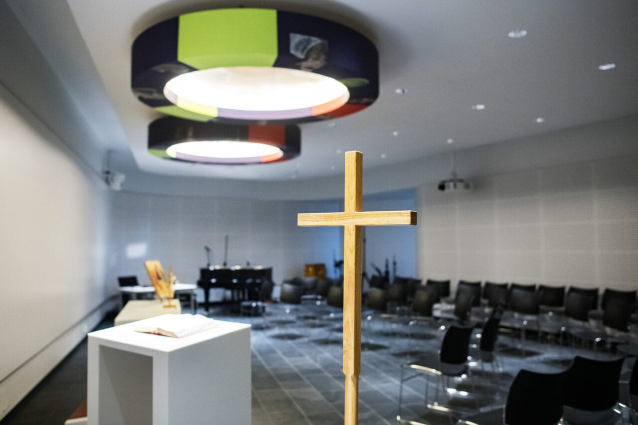 Kuvassa näkyy moderni kappeli tai rukoustila. Etualalla on vaalea puinen risti ja sen takana valkoinen pöytä, jonka päällä on avoin kirja. Taustalla on riveittäin mustia tuoleja ja musiikkivälineitä, kuten piano ja mikrofoneja.