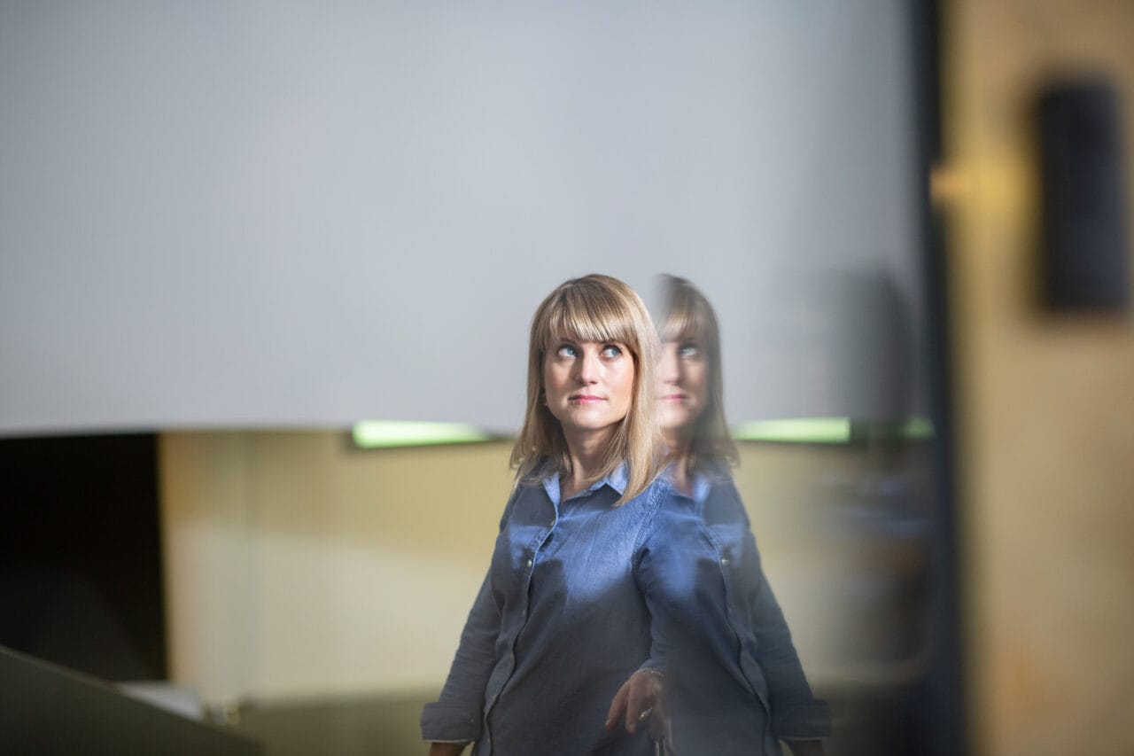 Nainen seisoo sisällä ja katsoo sivulle. Etualalla heijastava lasipinta, joka luo heijastuksen naisesta.