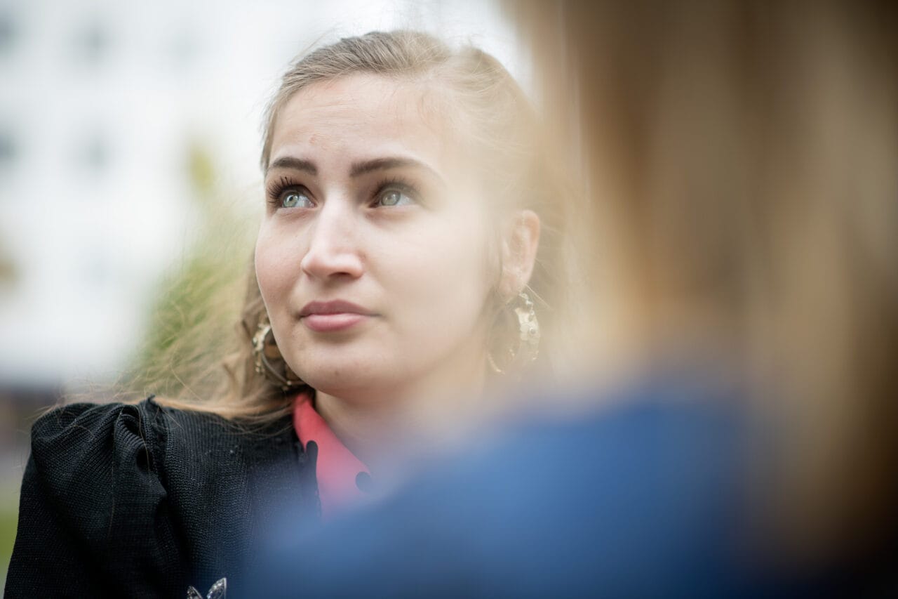 Nainen katsoo ulkona ylöspäin. Etualalla sumea hahmo ja taustalla vaalea rakennus ja puita.