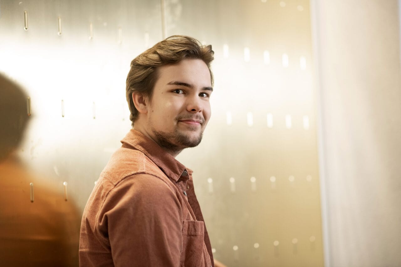 Tummahiuksinen mies seisoo seinän edustalla hymyillen.