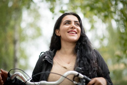 Nainen hymyilee ulkona ja katsoo sivulle. Yllään musta takki ja beige neule: Hänen kätensä ovat polkupyörän ohjaustangolla ja taustalla on vihreitä puita.