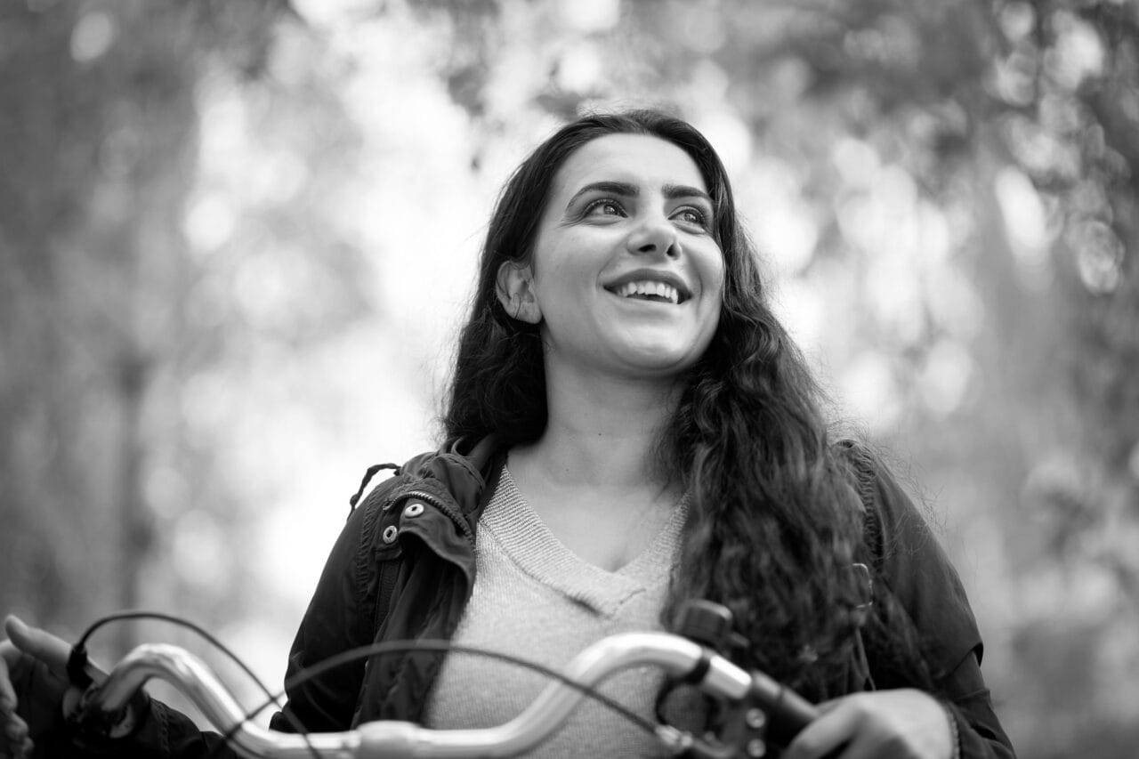Nainen hymyilee ulkona ja katsoo sivulle. Yllään musta takki ja beige neule. Hänen kätensä ovat polkupyörän ohjaustangolla ja taustalla on puita. Kuva on mustavalkoinen.