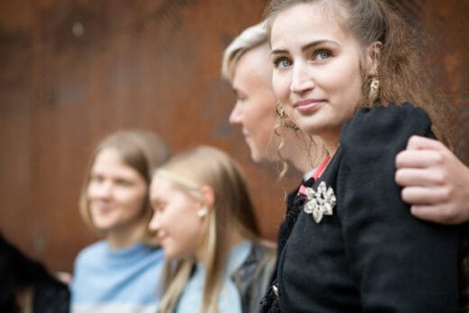 Etualalla nainen katsoo hieman ohi kameran. Hänen vieressään oleva henkilö pitää käsivarttaan naisen ympärillä. Taustalla muita henkilöitä ja ruskea seinä.