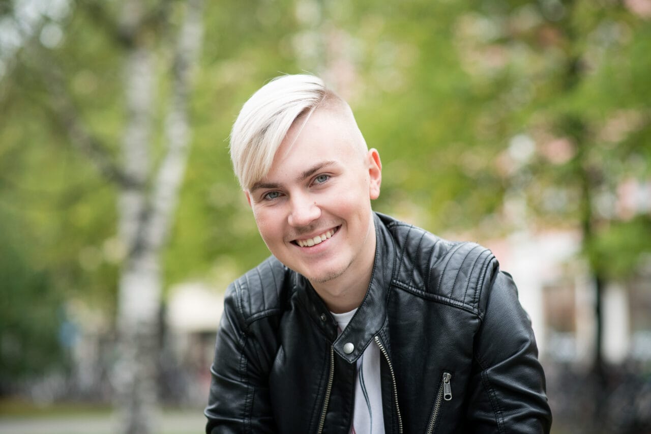 Mies hymyilee ulkona ja katsoo kameraan. Yllään musta nahkatakki, taustalla vihreitä puita.