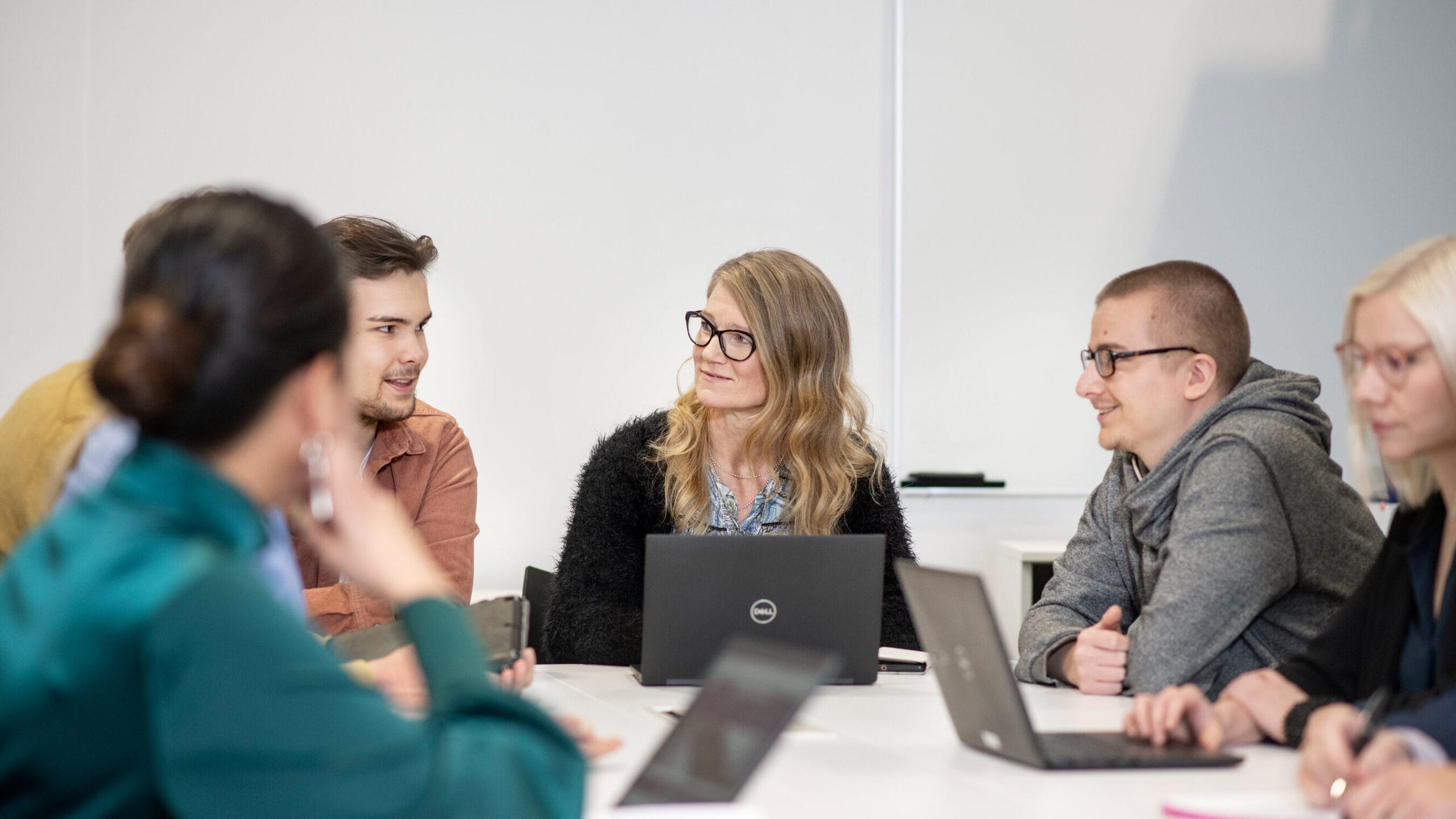 Viisi työntekijää saman pöydän ääressä ja avonaisia läppäreitä pöydällä.