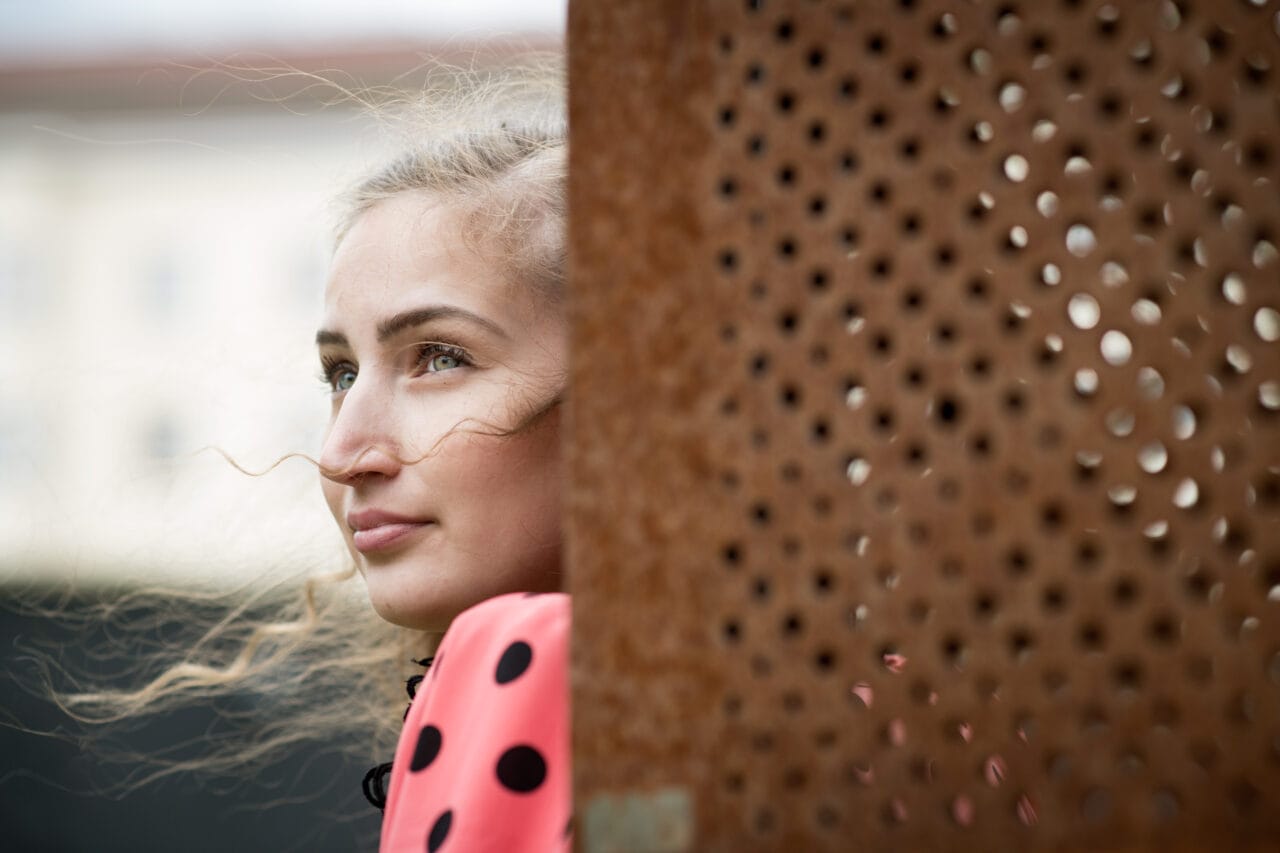 Nainen kurkistaa ulkona reiällisen ruosteenvärisen seinän takaa ja katsoo sivulle.