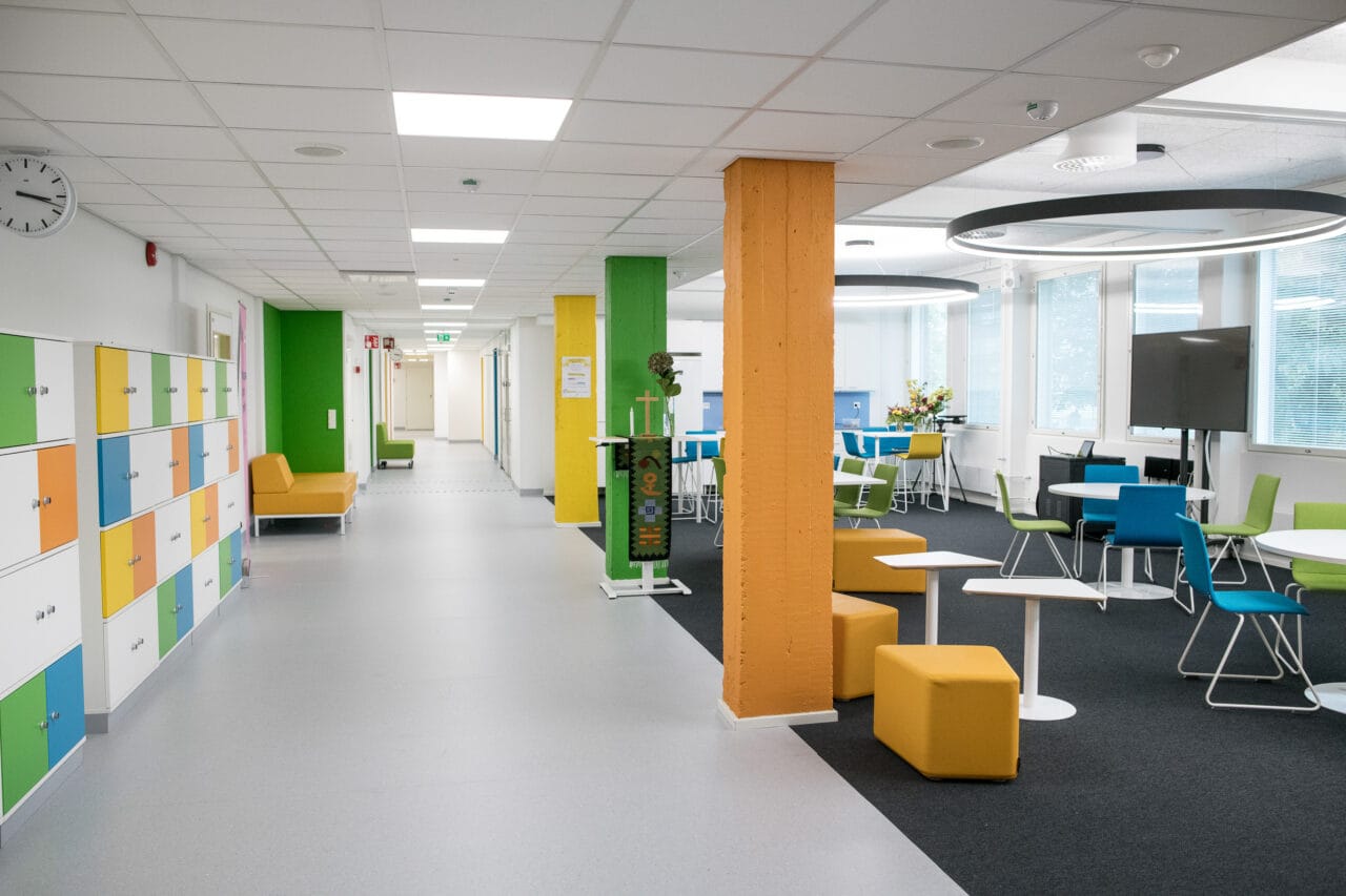 Oulun-kampuksen valoisa sisäkäytävä ja oleskelutila, jossa on värikkäitä kaappeja, pylväitä, pöytiä ja tuoleja sekä ikkunoita oikealla.