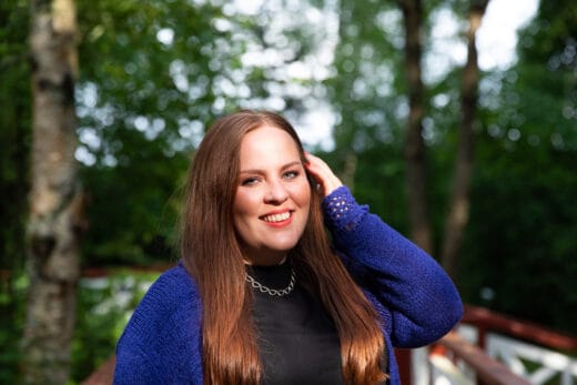 Ruskeahiuksinen nainen hymyilee kesäisessä luonnossa.