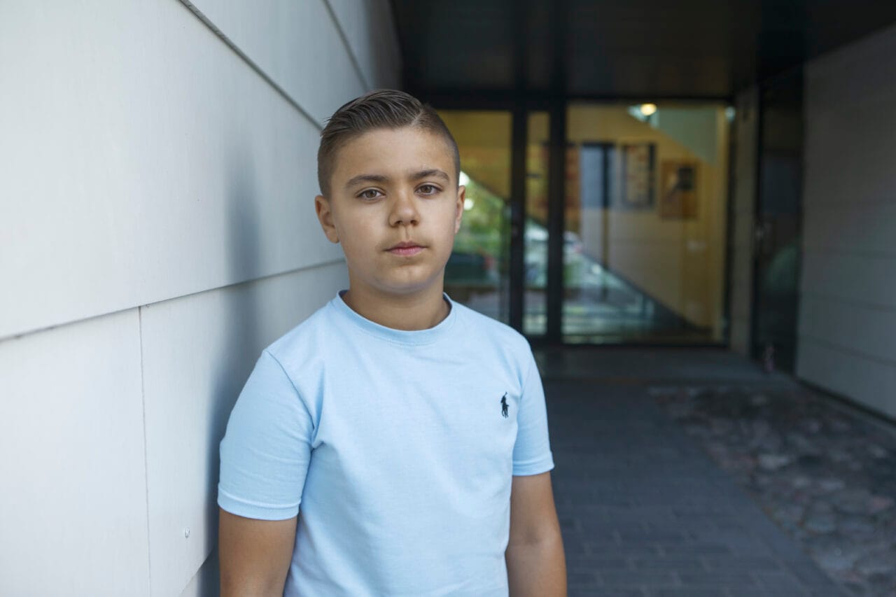 Poika seisoo ulkona rakennuksen seinän vieressä ja katsoo kameraan, yllään vaaleansininen t-paita.