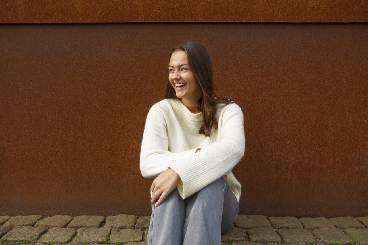 Tyttö istuu maassa ruskean seinän edessä ja nauraa katsoen sivulle. Yllään vaalea neule ja harmaat housut.