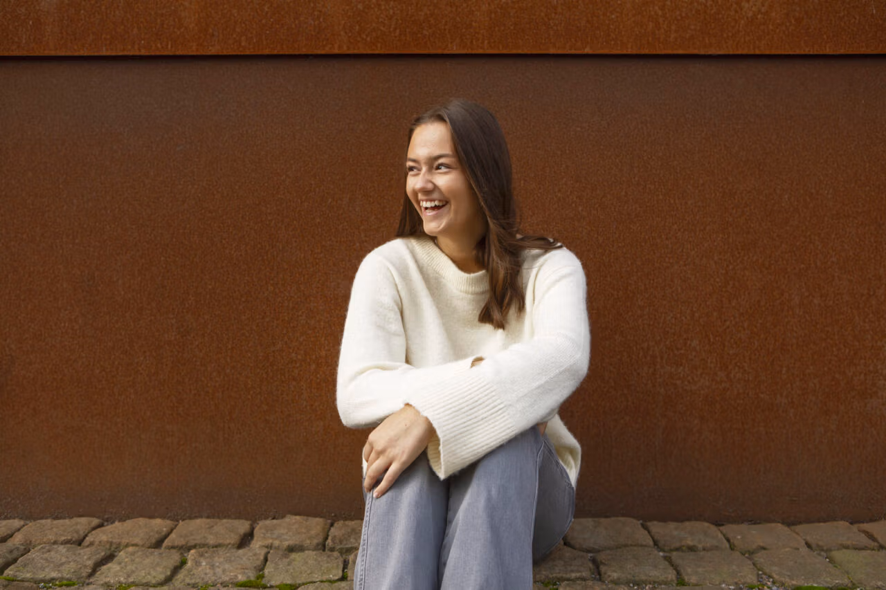 Tyttö istuu maassa ruskean seinän edessä ja nauraa katsoen sivulle. Yllään vaalea neule ja harmaat housut.