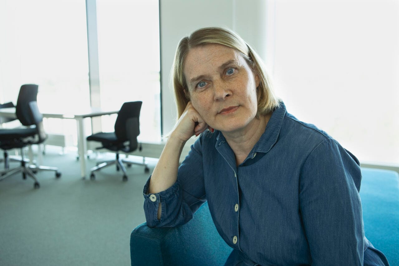 Nainen istuu sisällä ja katsoo kameraan, yllään sininen paita, taustalla työpöytiä ja tuoleja ikkunan edessä.