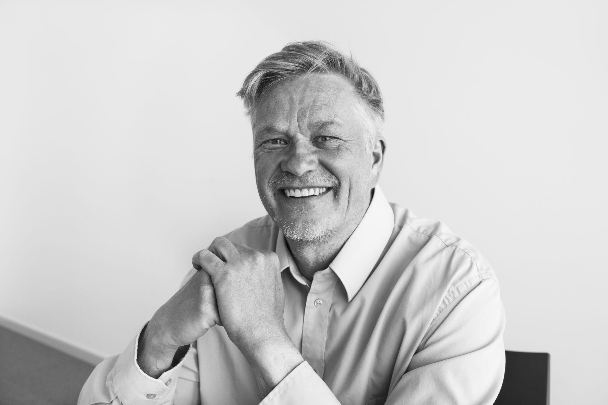 Mies istuu sisällä ja hymyilee kameraan, yllään vaalea kauluspaita, taustalla vaalea seinä ja tuoli. Kuva on mustavalkoinen.