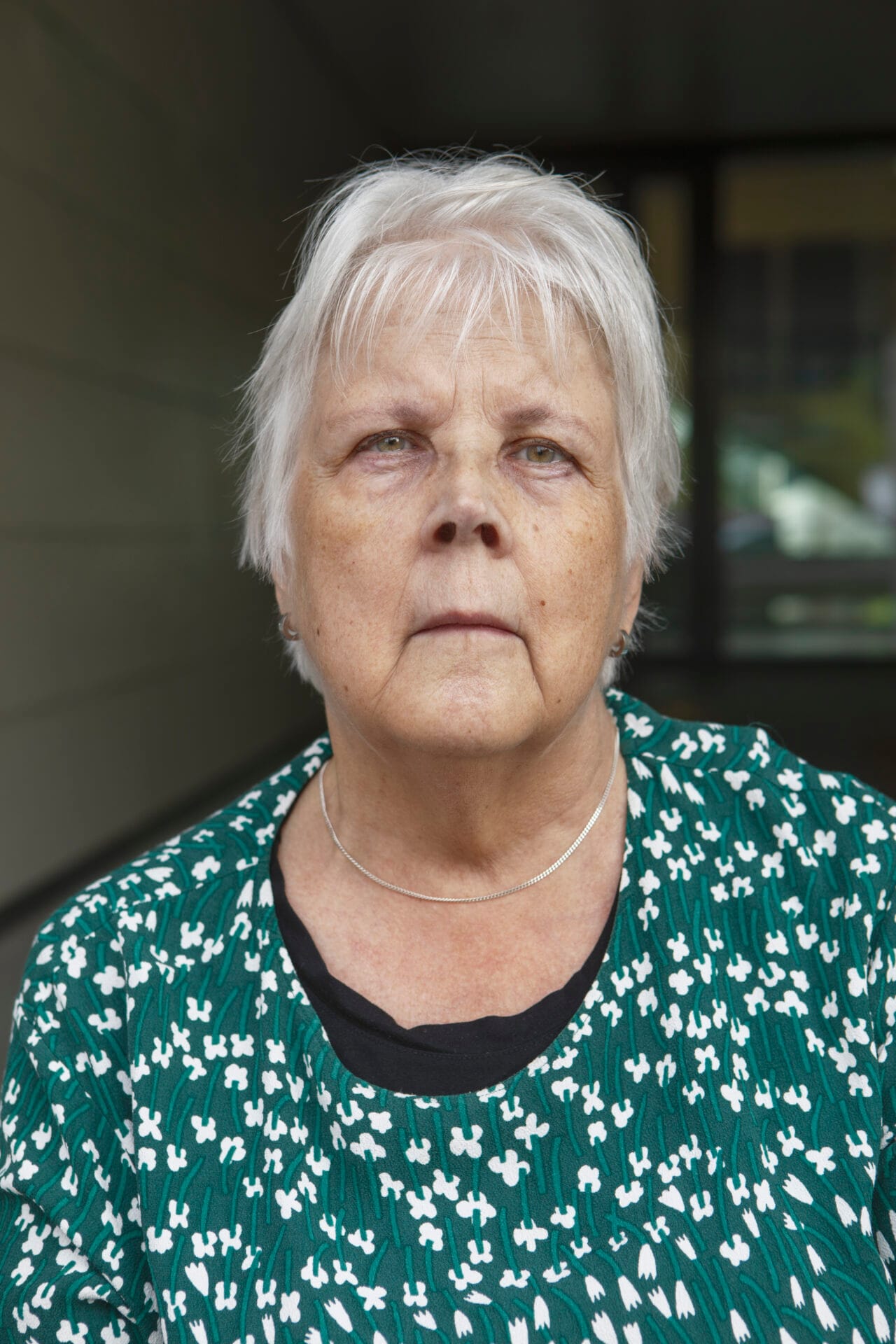 Iäkäs nainen vihreässä kuviollisessa paidassa katsoo vakavana kameraan.