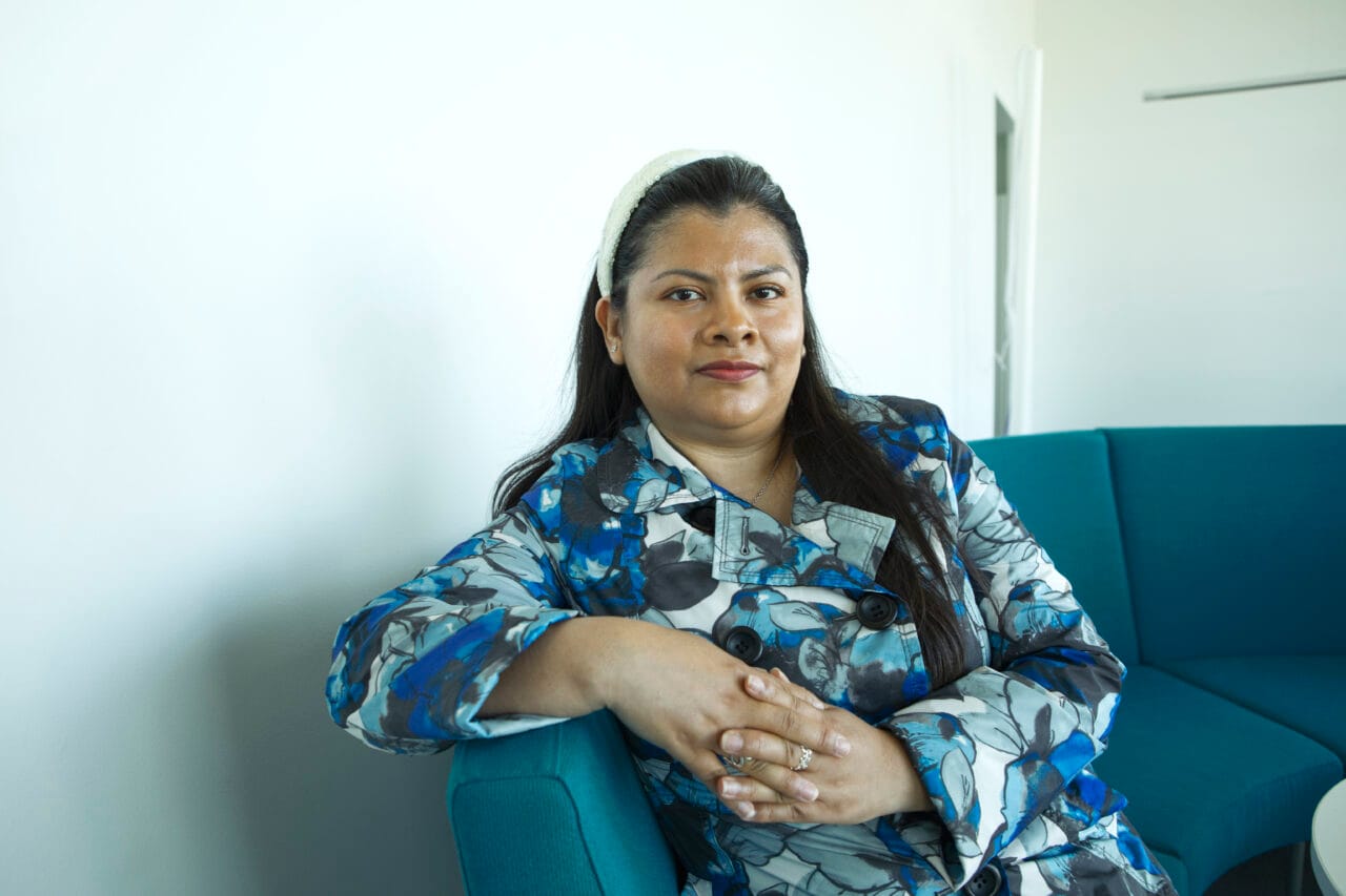 Nainen istuu sisällä turkoosilla sohvalla ja katsoo kameraan. Yllään sinikuvioinen takki, taustalla vaalea seinä.