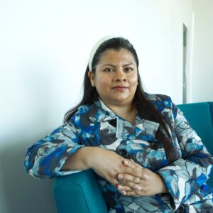 Nainen istuu sisällä turkoosilla sohvalla ja katsoo kameraan. Yllään sinikuvioinen takki, taustalla vaalea seinä.