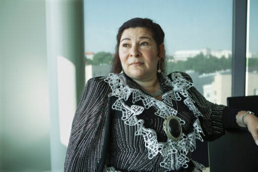 Nainen seisoo sisällä ikkunan edessä ja katsoo sivulle. Yllään romaninaisen puku. Taustalla kaupunkinäkymä.