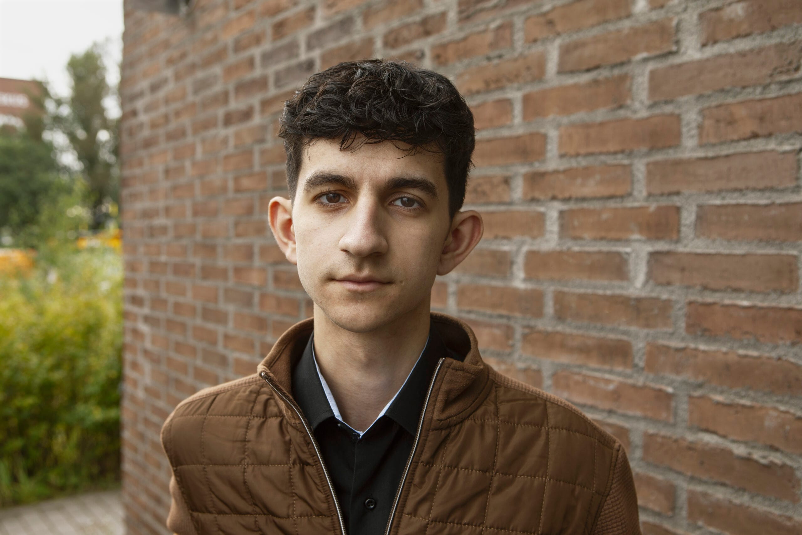 Nuori mies seisoo tiiliseinän edessä ja katsoo suoraan kameraan.