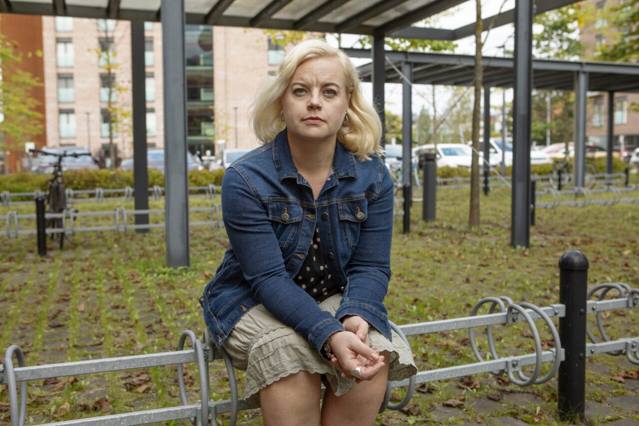 Nainen istuu ulkona pyörätelineellä ja katsoo kameraan. Taustalla katos, puita ja kerrostaloja.