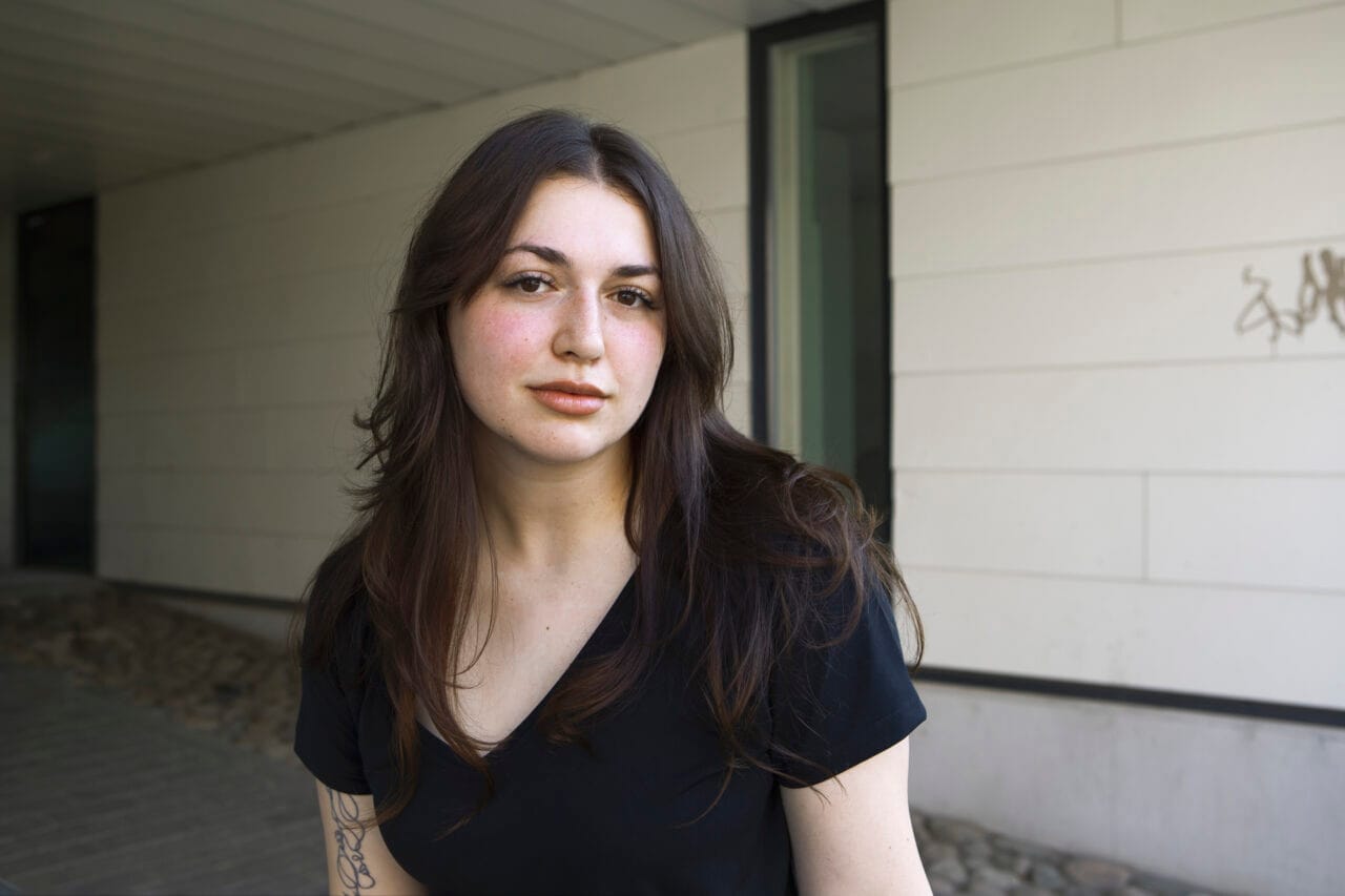 Nuori nainen seisoo ulkona vaalean rakennuksen seinän edessä ja katsoo kameraan.