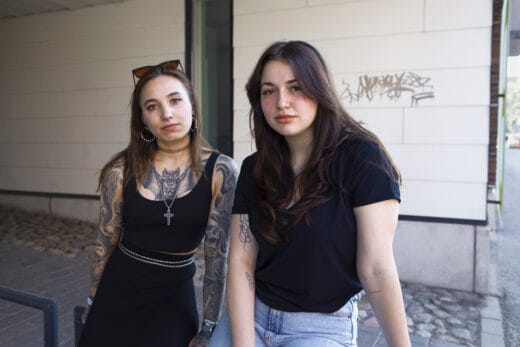 Kaksi ihmistä seisoo vierekkäin ulkona rakennuksen seinän edessä ja katsoo kameraan. Toisella on tatuoidut käsivarret. Taustalla vaalea seinä ja pihakivetys.