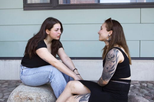 Kaksi ihmistä istuu vastakkain kivellä ja juttelee. Taustalla vaalea rakennuksen seinä.