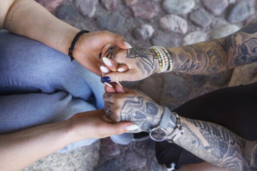 Ylhäältä kuvattu kuva kahden henkilön käsistä, jotka pitävät toisiaan kädestä. Toisella on tatuoidut käsivarret ja rannekoruja. Taustalla kivetys.