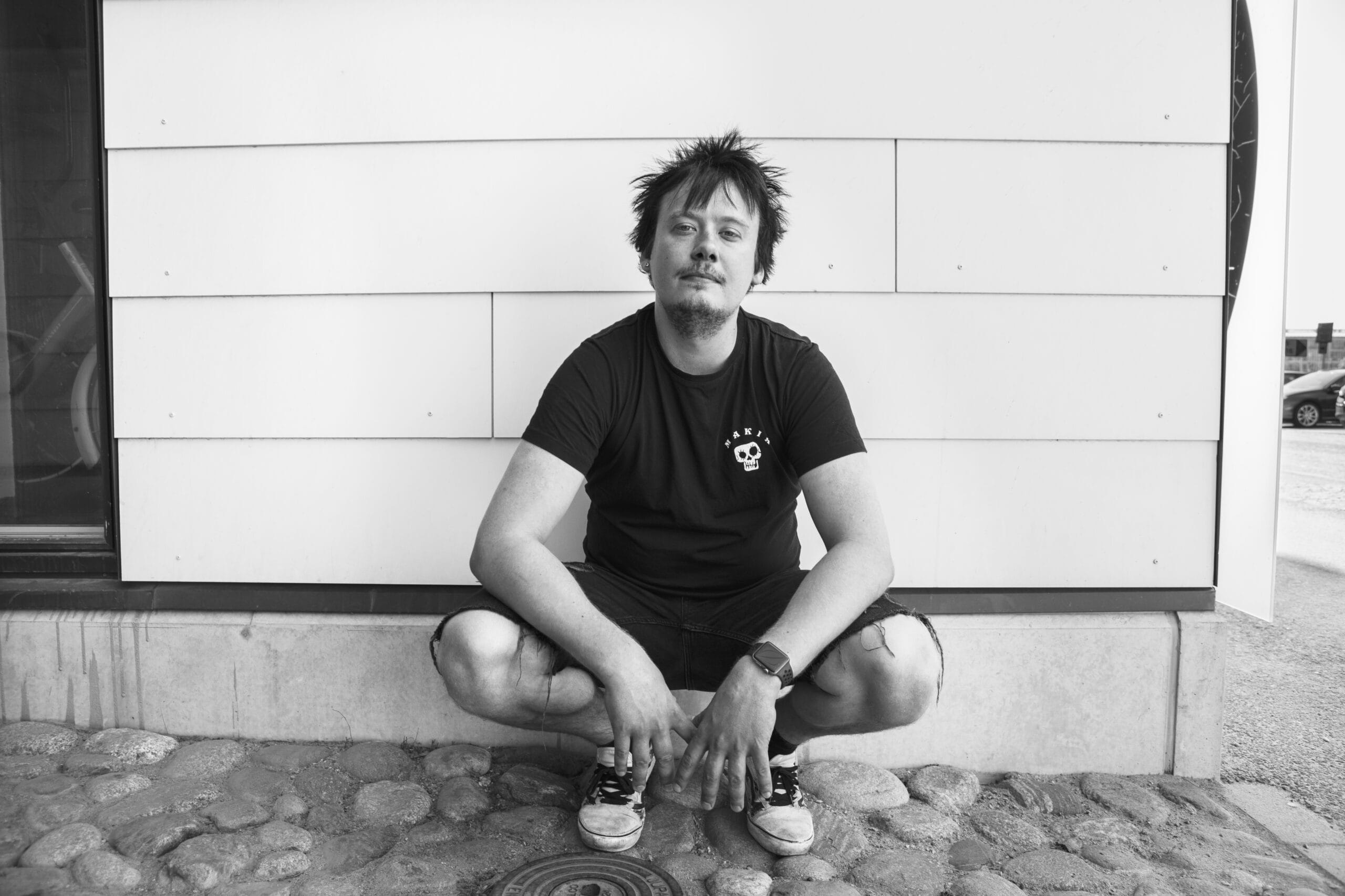 Nuori mies on kyykyssä seinän edessä ja katsoo kameraan. Kuva on mustavalkoinen.