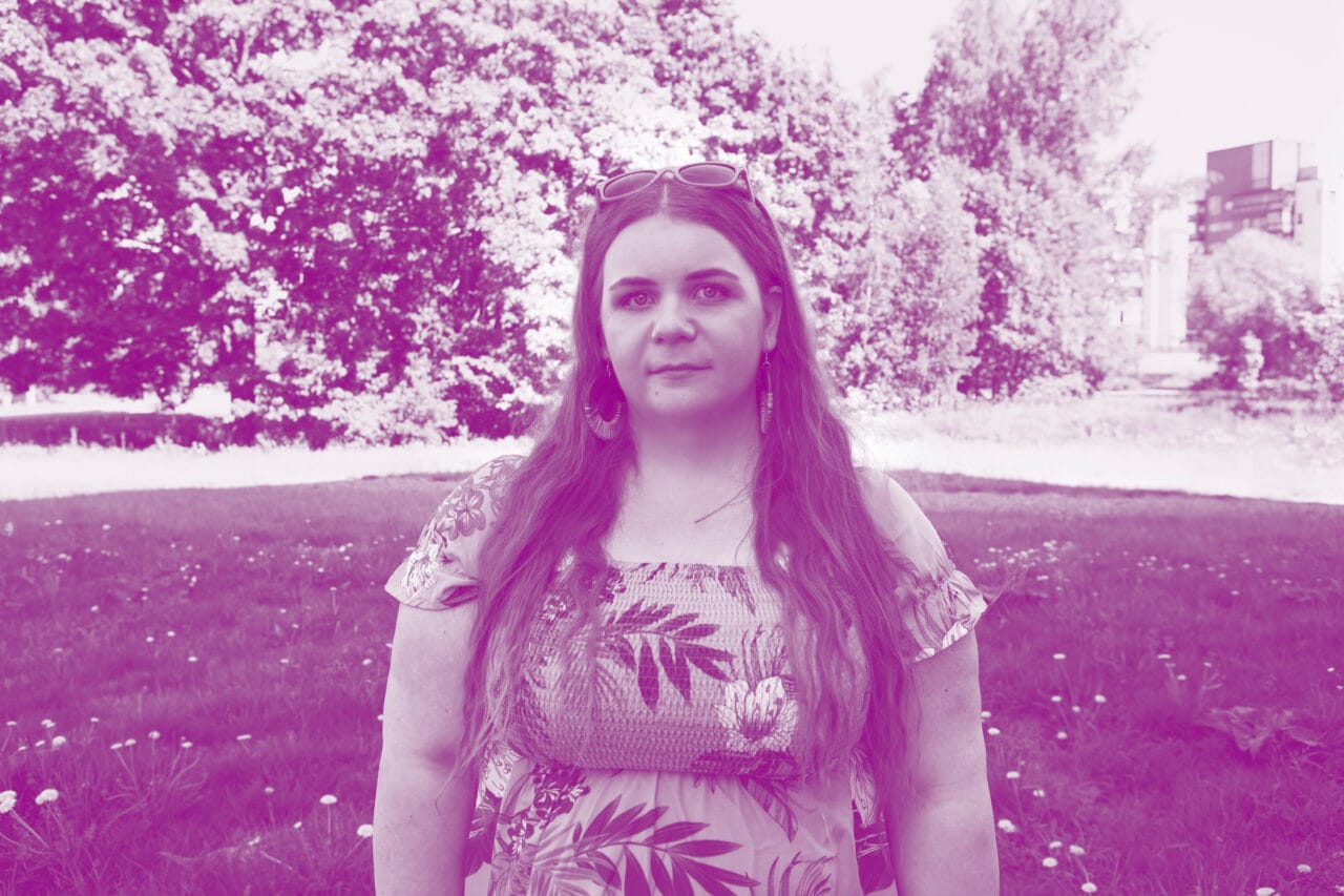 Kukkamekkoinen nainen seisoo nurmikolla. Kuva on sävytetty violetiksi.