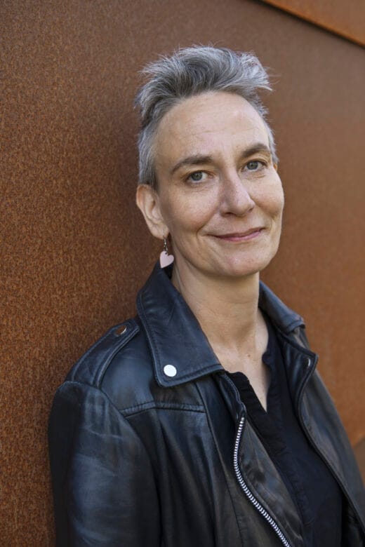 Nainen katsoo kameraan ja hymyilee. Naisella lyhyt harmaa tukka ja korvakorut.