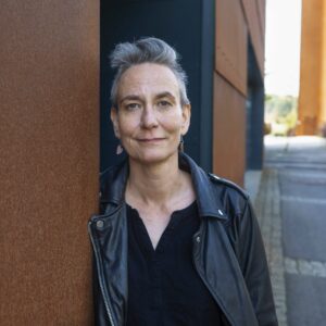 Nainen, jolla harmaa lyhyt tukka, katsoo kameraan, hymyilee ja nojaa seinään.